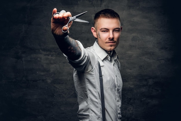 Portrait of tattooed male barber holds sharp scissors and a blade.