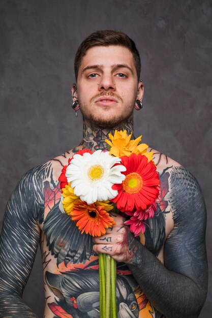 Portrait of a tattooed man with piercing in the ears and nose holding colorful gerbera flowers in hand