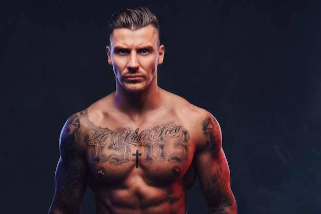 Portrait of a tattooed muscular shirtless man with stylish hair posing at the camera on a dark background.