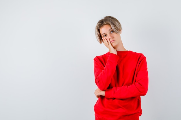 Portrait of teen boy keeping hand on cheek in red sweater and looking fatigued front view