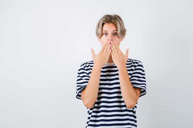 Portrait of teen boy keeping hands on mouth in t-shirt and looking shocked front view