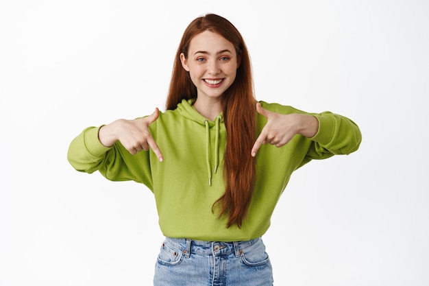 Portrait of teen ginger girl smiling white teeth, pointing fingers down at promo discount, show advertisement, standing against white background.