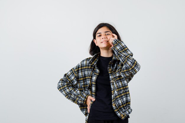 Портрет девочки-подростка, держащей палец на висках, держащей руку на талии в повседневной рубашке и выглядящей жизнерадостной, вид спереди