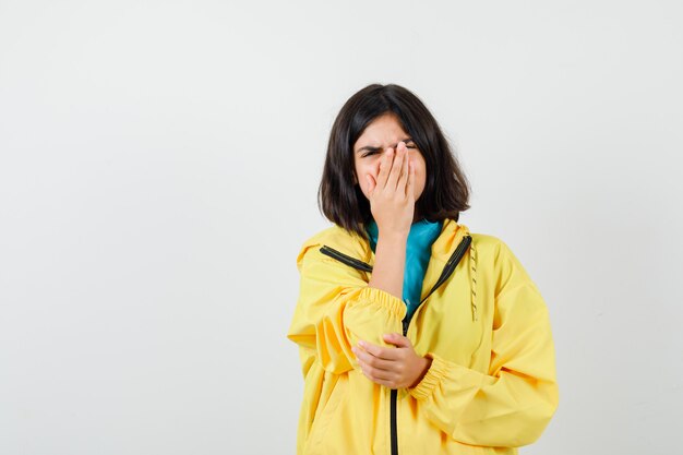 Portrait of teen girl holding hand on mouth in yellow jacket and looking sorrowful front view