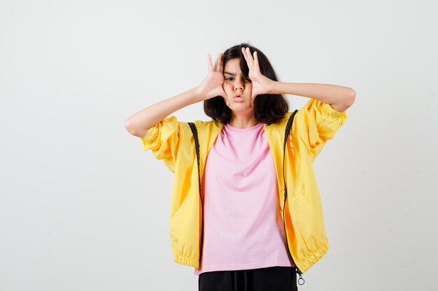 Портрет девушки-подростка, держащей руки по обеим сторонам лица в футболке, куртке и раздраженной взглядом спереди