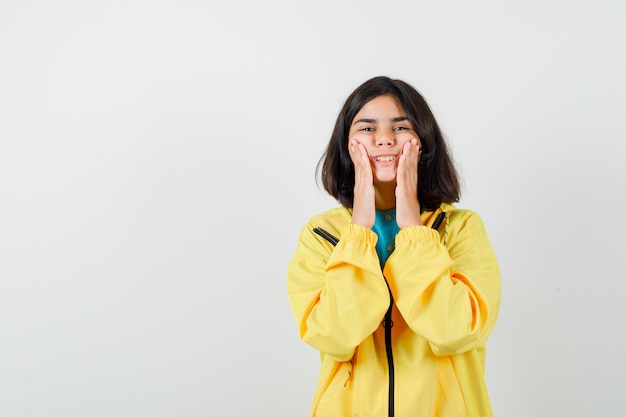 Portrait of teen girl pretending to rub skin cream on cheeks in yellow jacket and looking careful front view