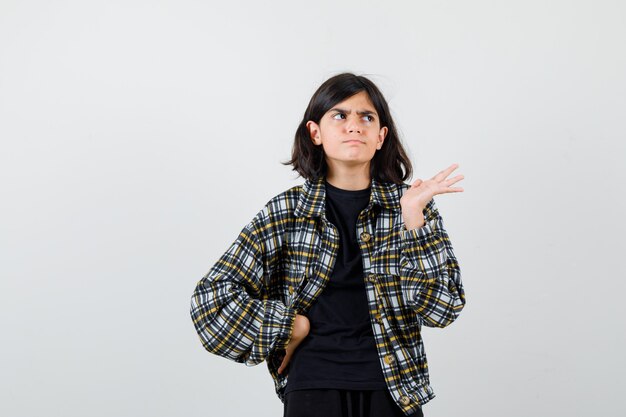 Portrait of teen girl spreading palm aside, holding hand on waist in casual shirt and looking indecisive front view