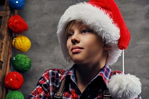 Portrait of teenage boy dressed in Santa's hat and a plaid shirt on grey background with colorful illumination.