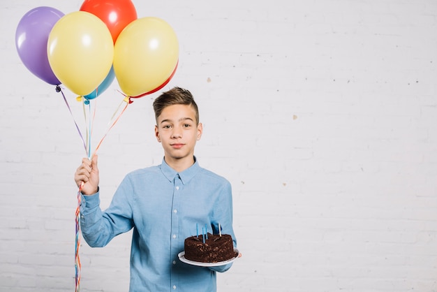 Free photo portrait of teenage boy holding balloons and birthday cake against wall