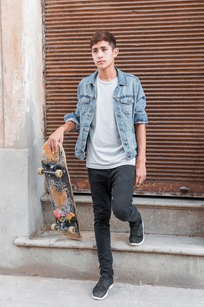Портрет подростка, стоящего перед закрытым сайдингом из гофрированного железа, держащего скейтборд
