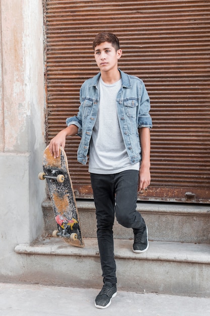 Portrait of teenage boy standing in front of closed corrugated iron siding holding skateboard