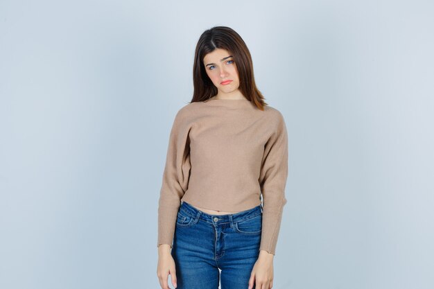 Portrait of teenage girl curving lower lip in sweater, jeans and looking disappointed front view