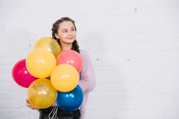 Portrait of a teenage girl holding balloons in hand standing against white wall