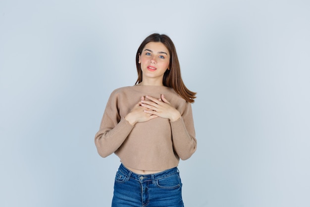 Portrait of teenage girl keeping hands on chest in sweater, jeans and looking grateful front view