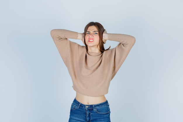 Free photo portrait of teenage girl keeping hands on ears, clenching teeth in sweater, jeans and looking nervous front view