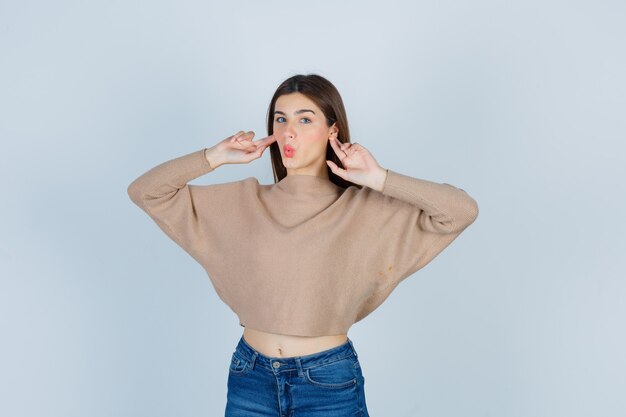 Portrait of teenage girl plugging ears with fingers, pouting lips in sweater, jeans and looking surprised front view