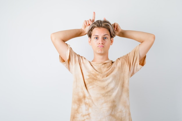 Free photo portrait of teenager boy keeping fingers over head as bull horns in t-shirt and looking sulky front view