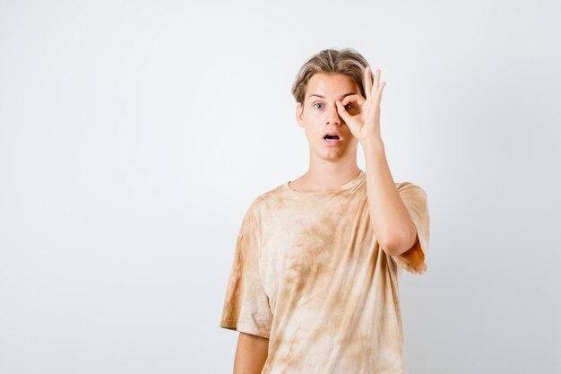 Portrait of teenager boy showing ok sign on eye in t-shirt and looking shocked front view