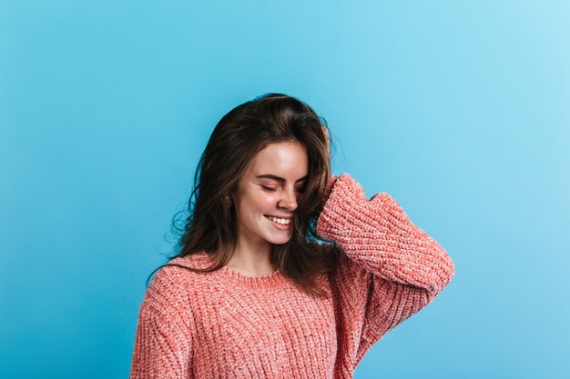 Portrait of teenager girl in pink sweater. Model smiles with closed eyes on blue wall.