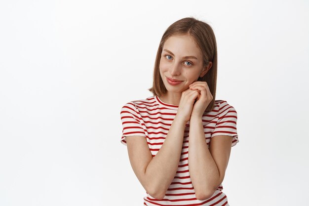 Portrait of tender and beautiful young woman with blond hair, no makeup clean face, admire smth, looking coquettish and lovely at front, dreaming, standing in t-shirt against white wall.