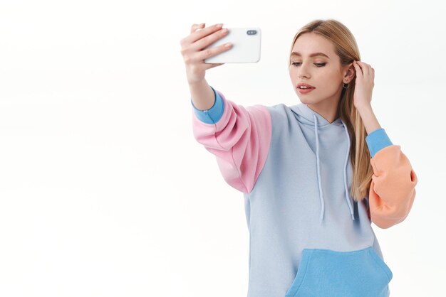 Portrait of tender feminine beautiful blonde girl put hair strand behind ear looking down sensual taking selfie on smartphone apply photo filter using mobile app standing white background