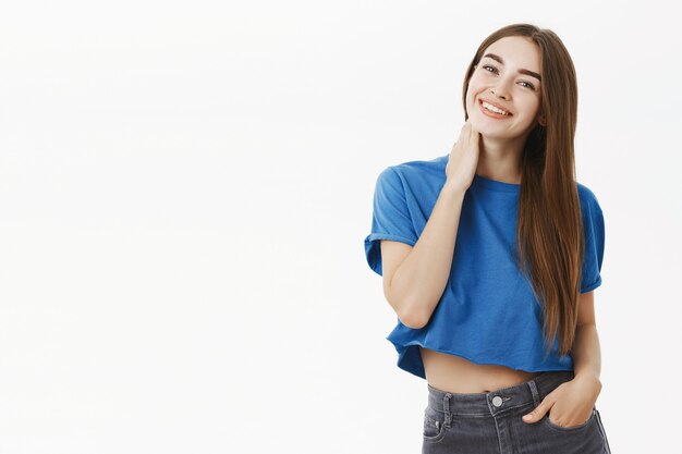 Portrait of tender and feminine cute female in cropped blue t-shirt touching neck shy and flirty tilting head and smiling broadly holding hand in pocket being delighted with attention