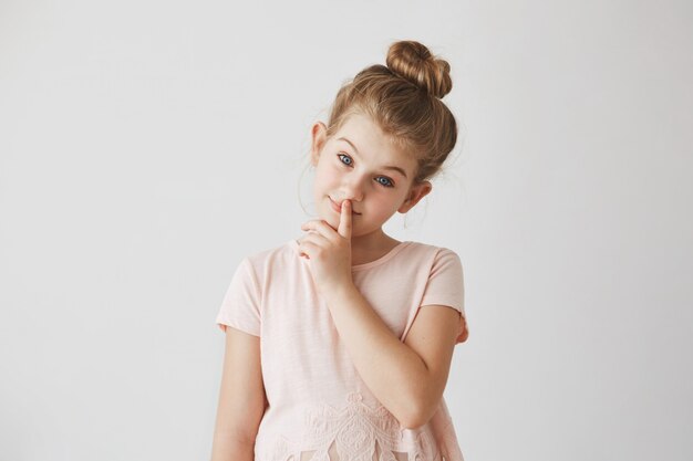 Portrait of tender little girl with blond hair in bun hairstyle , holding finger on lips and smiling,  with calm face expression.