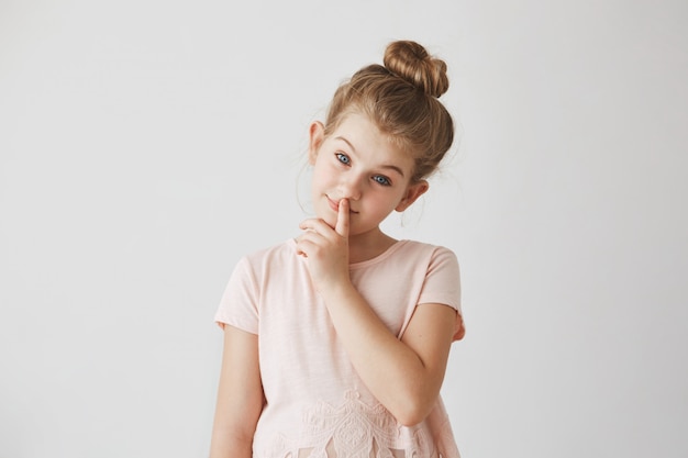 Портрет нежная маленькая девочка со светлыми волосами в булочке прическа, держа палец на губах и улыбается, с спокойным выражением лица.