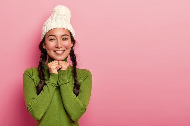 Portrait of tender lovely Asian woman has rouge cheeks, pleasant smile, keeps hands under chin, looks happily directly at camera, wears knitted hat