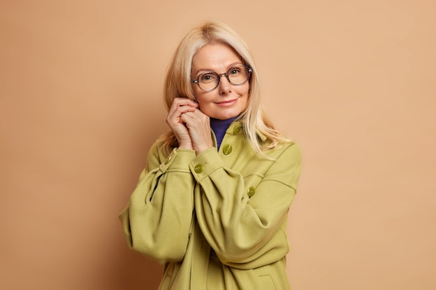 Portrait of tender middle aged woman keeps hands near face looks directly at camera with calm expression