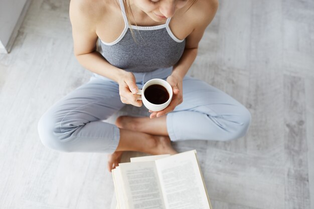 Portrait of tender young beautiful woman reading book holding cup of coffee sitting on floor over white wall. 