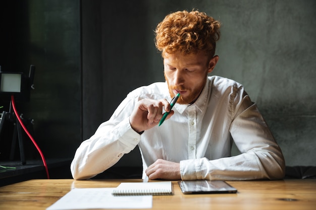 Portrait of thinking young readhead man in white shirt, sitting at wooden table with pen in his mouth