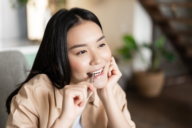 Portrait of thoughtful asian girl biting pen and smiling thinking searching inspiration doing homewo...