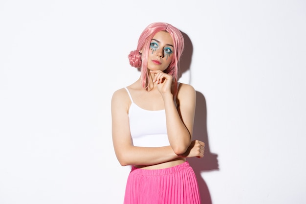 Portrait of thoughtful attractive female model with pink anime wig and bright makeup, looking at upper left corner banner and thinking, making choice, standing.