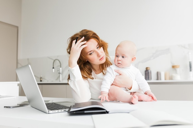 Free photo portrait of thoughtful business woman holding her little baby while sitting at the table and working