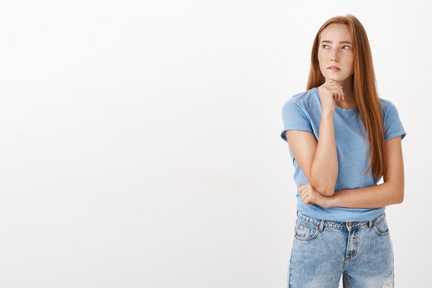 Portrait of thoughtful concentrated cute and feminine woman with long red hair and freckles turning left squinting supporting head with fist while thinking making decision or being doubtful