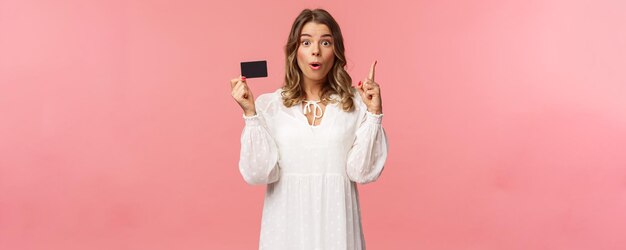 Portrait of thoughtful cute blond girl in white dress have and excellent idea holding credit card say wow eureka raise index finger have suggestion know where find needed product