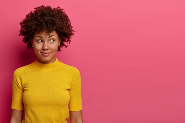 Portrait of thoughtful dreamy woman has gentle smile, looks aside, thinks over something, wears casual yellow t shirt, satisfied with pleasant thoughts, poses over rosy wall, blank space aside