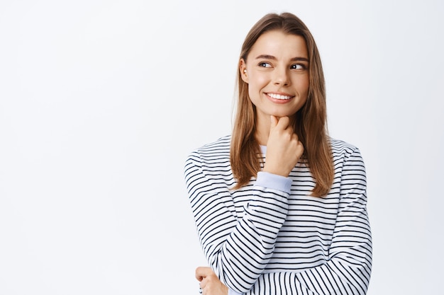 Portrait of thoughtful girl making assumption, having interesting ideas, looking left at copy space logo and smiling, pondering, standing over white wall