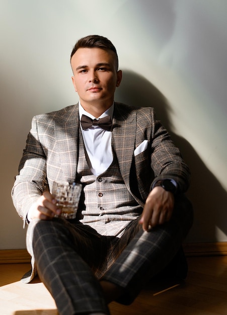 Portrait of thoughtful and handsome groom wearing in brown plaid suit with bow tie sitting on floor with glass of alcohol beverage on white background seriously looking at camera