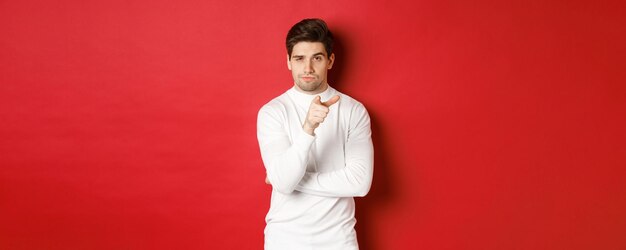 흰색 스웨터를 입은 사려 깊은 잘생긴 남자의 초상화, 카메라를 가리키고 선택을 하고 빨간색 배경에 서 있습니다.