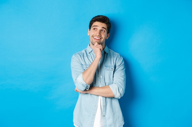 Portrait of thoughtful handsome man with beard, standing in casual outfit, looking at upper left corner and smiling, imaging or dreaming about something, standing over blue background.