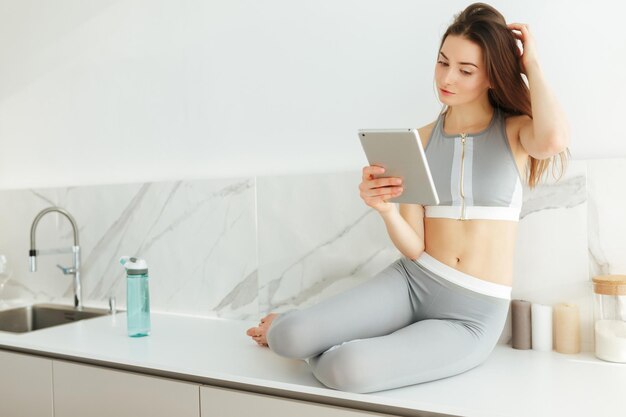 Portrait of thoughtful lady in sporty top and leggings sitting on kitchen table with tablet in hand