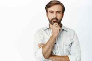 Free photo portrait of thoughtful mature man looking serious at camera touching beard and thinking making choice white background