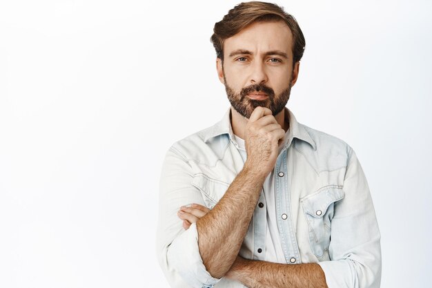 Портрет вдумчивого зрелого мужчины, серьезно смотрящего в камеру, трогающего бороду и думающего, делающего выбор на белом фоне