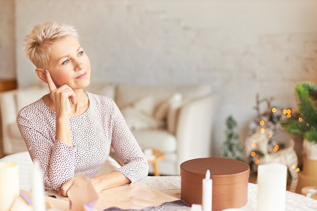 Portrait of thoughtful middle aged female in elegant dress sitting at table in room decorated with garland having pensive look, holding index finger on her head, thinking how to wrap gifts for family