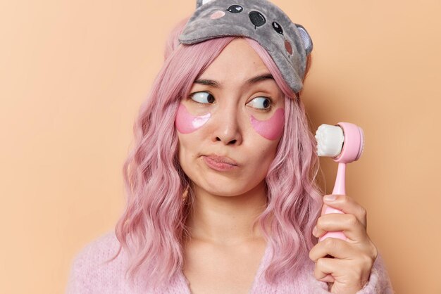 Portrait of thoughtful pink haired Asian woman looks at face massager and thinks how to use it applies beauty patches under eyes for skin treatment wears sleepmask isolated over brown background