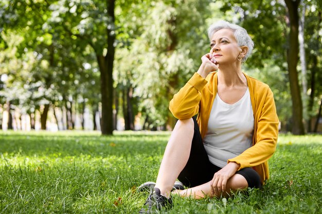 Portrait of thoughtful retired woman in running shoes sitting comfortably on green grass, holding hand under her chin, watching people walk in park with pensive facial expression, feeling relaxed