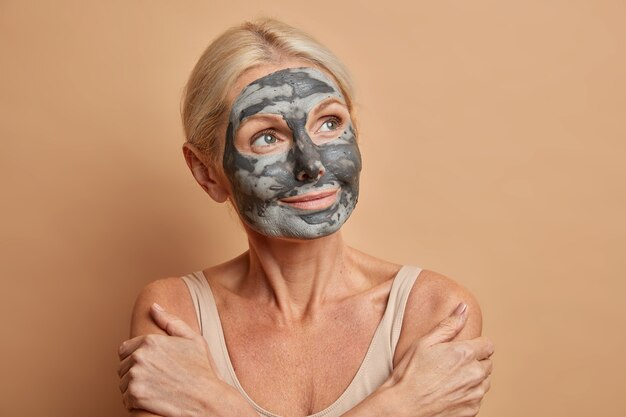 Portrait of thoughtful senior Caucasian woman crosses arms and touches shoulders gently wears homemade facial mask at home concentrated away isolated on beige wall