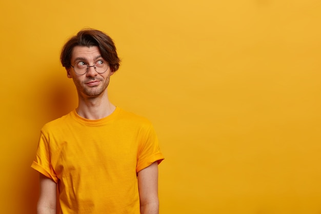 Portrait of thoughtful serious man concentrated somewhere aside, thinks how better to act, wears big round spectacles and casual t shirt, isolated on yellow wall, empty space. Monochrome